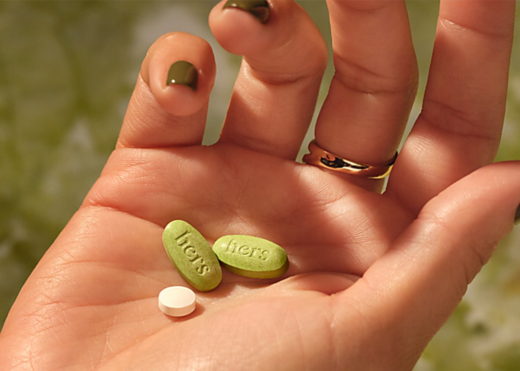 A hand holding two green oval pills with "hers" embossed on them and one small white round pill.