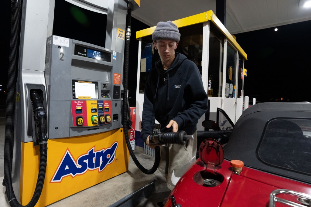 A person in a gray beanie and a black hoodie filling the tank of their red Mazda Miata at an Astro gas station.