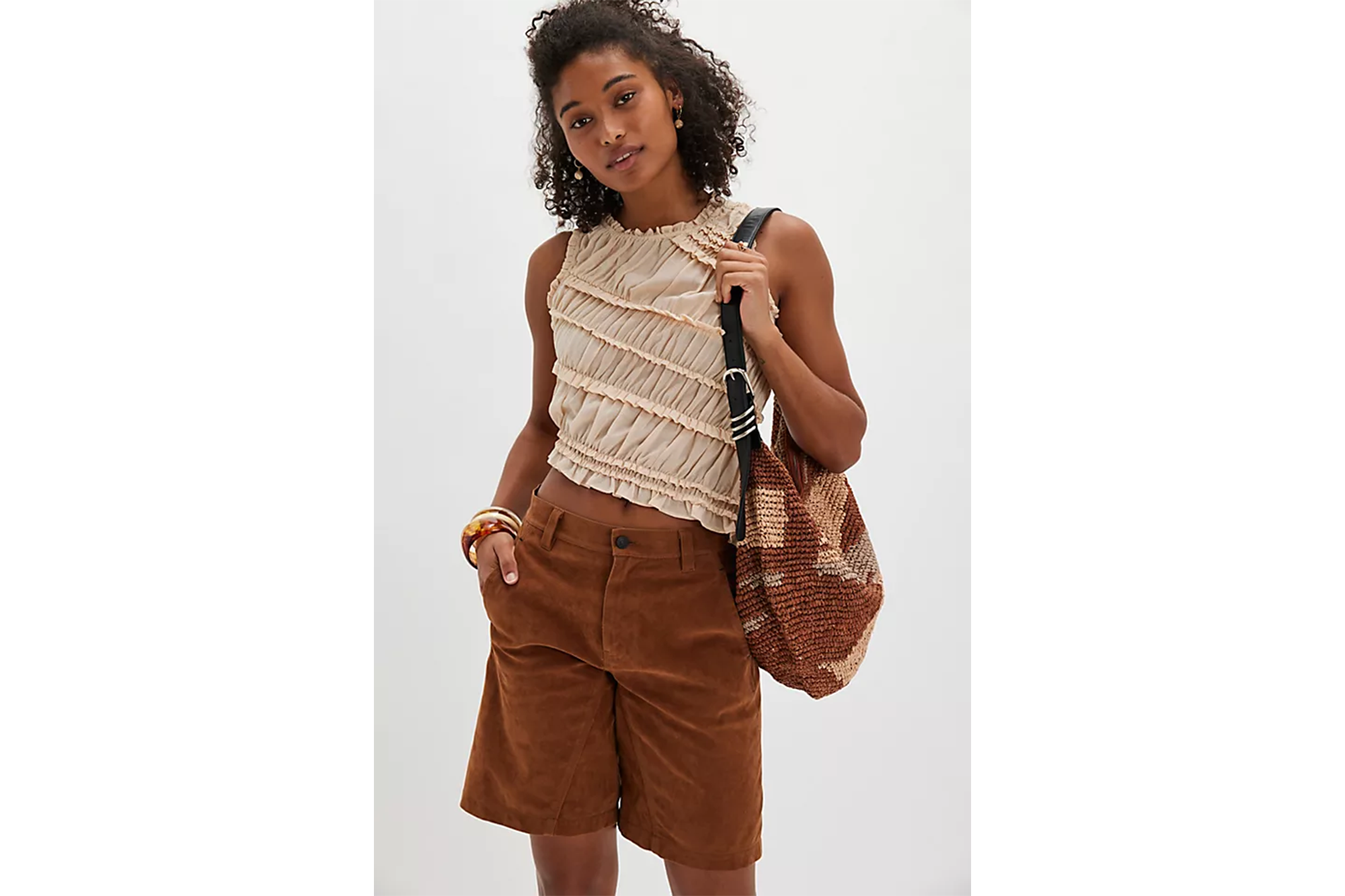 A model wearing a ruffled, beige sleeveless top and brown shorts, with a knit bag over her shoulder.