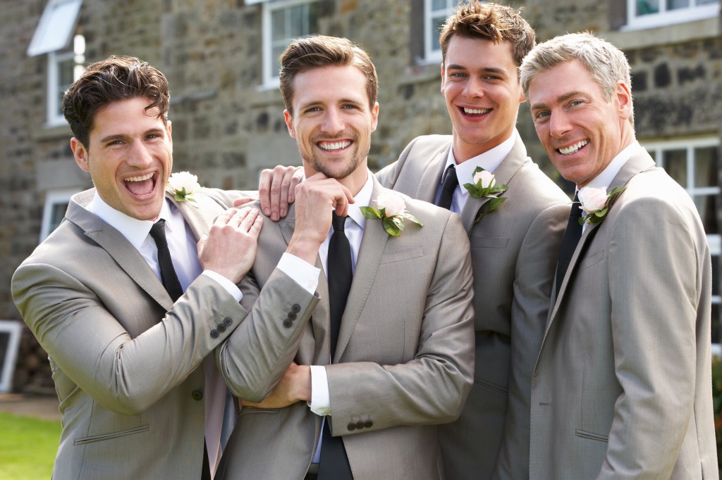 Four happy men, including a groom, best man, and two groomsmen, smiling at the camera outdoors.