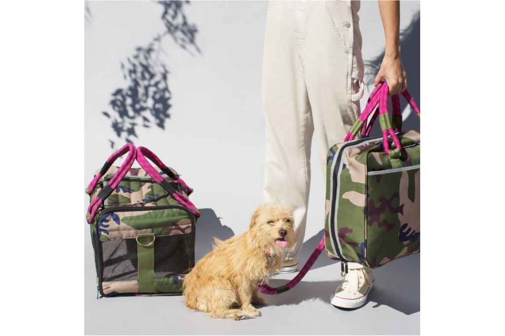 A small yellow dog in front of a woman's feet carrying a camo and pink bag and a matching dog carrier next to them 