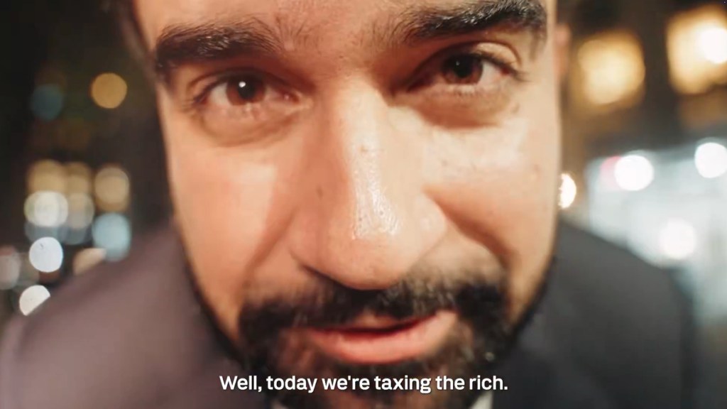 Close-up of a man with dark hair and a beard, looking intensely at the camera, with the subtitle "Well, today we're taxing the rich."