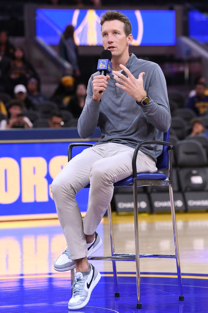 A man in a grey shirt and khaki pants sits on a tall stool, holding a microphone with a Golden State Warriors logo.