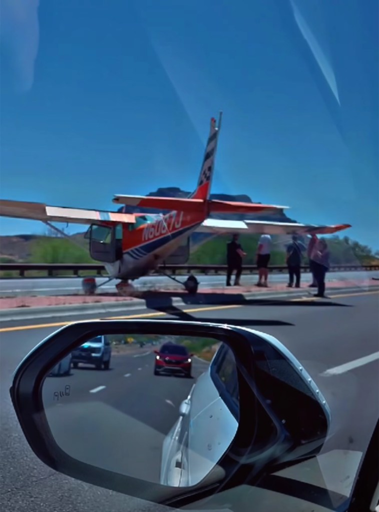 A small orange and white plane landed on a highway in Arizona, with people standing around it, seen from a car's perspective with the side mirror reflecting other vehicles.