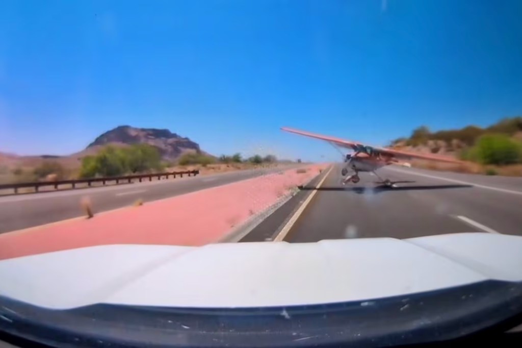 A small plane lands on a desert highway, viewed from a car's dashboard.