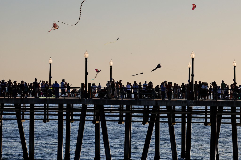 Silhouetted crowd on a pier with several kites flying in the sunset sky.