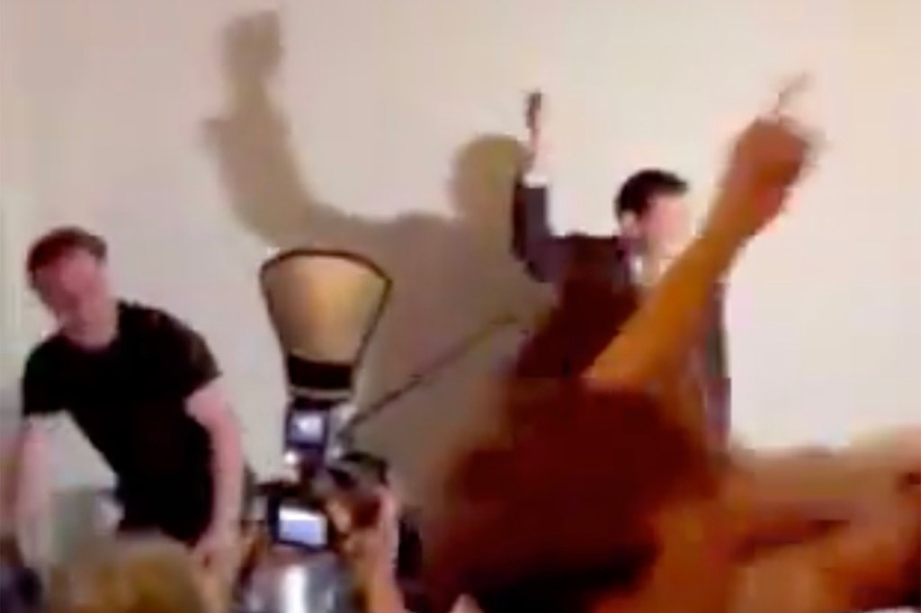 A blurry image showing a man in a suit raising his arms behind a DJ booth, with a shadow of another person on the wall behind him.