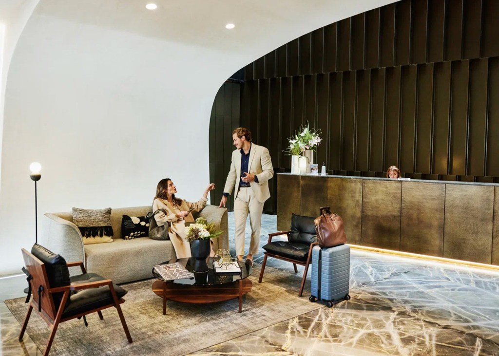 A man and woman talking in a modern hotel lobby, with a suitcase nearby.