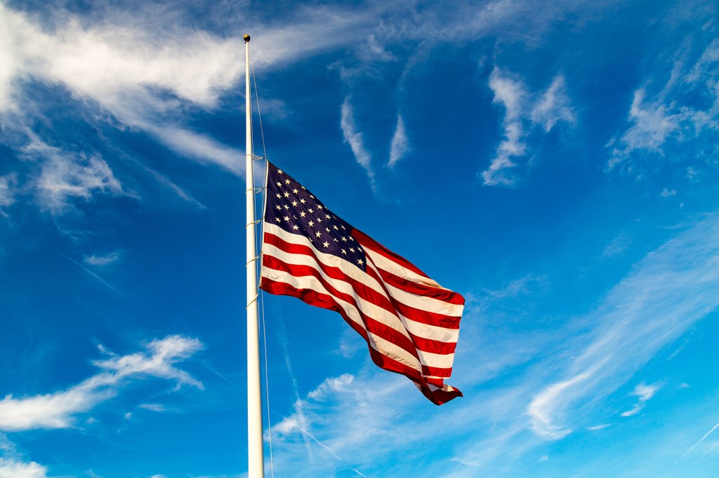 USA flag flying at half-mast against a blue sky with white clouds.